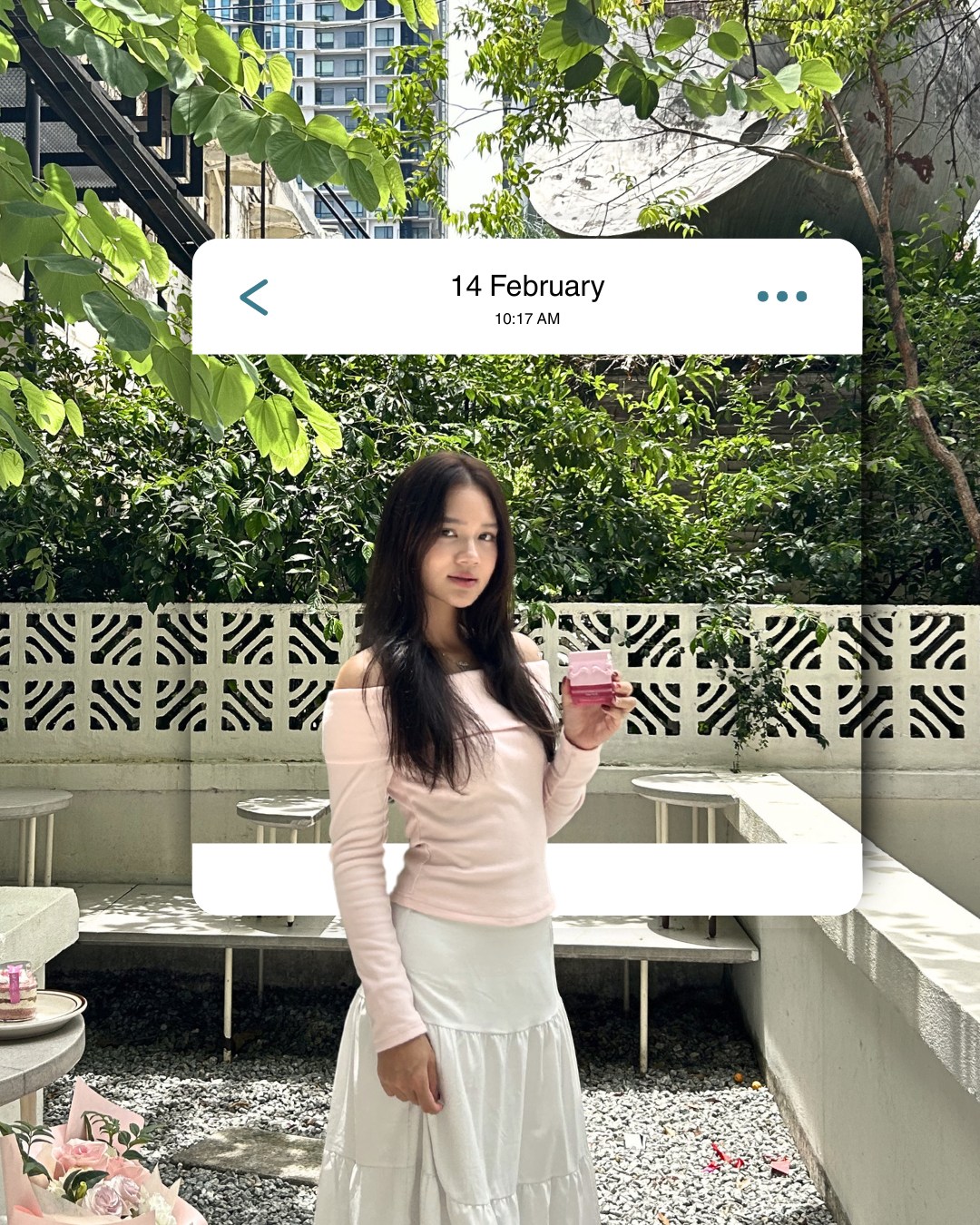 A woman in a pink off-shoulder top and white skirt holding a small pink bottle in a garden courtyard, inside a '14 February' message card (slide 4 of 4)