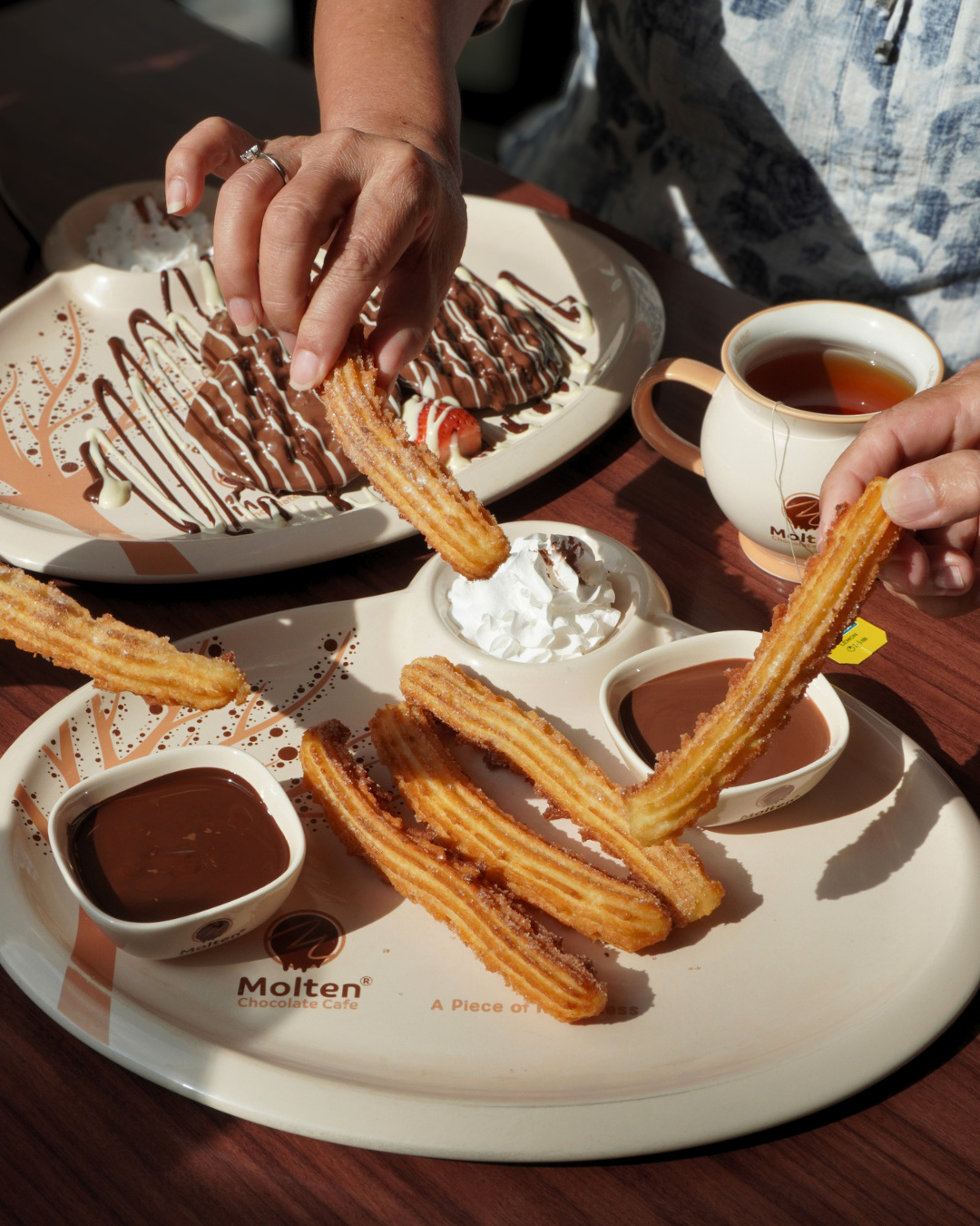Hands dipping churros into chocolate sauce beside a chocolate crepe and a cup of tea (slide 1 of 4)