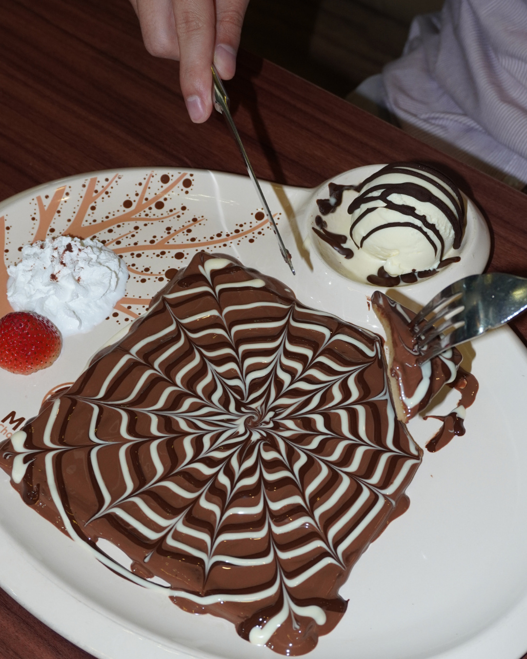 A swirl-patterned chocolate crepe with ice cream, whipped cream and a strawberry, being cut with a knife and fork (slide 2 of 4)