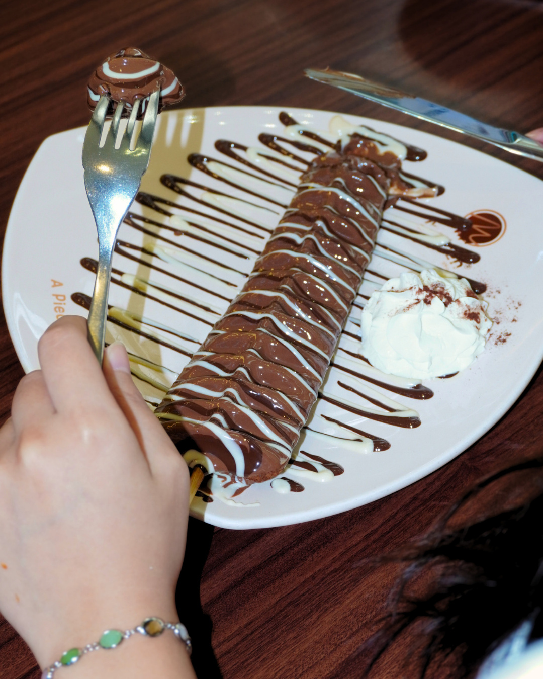 A chocolate crepe roll drizzled with white chocolate on a plate, with a fork lifting a bite (slide 3 of 4)